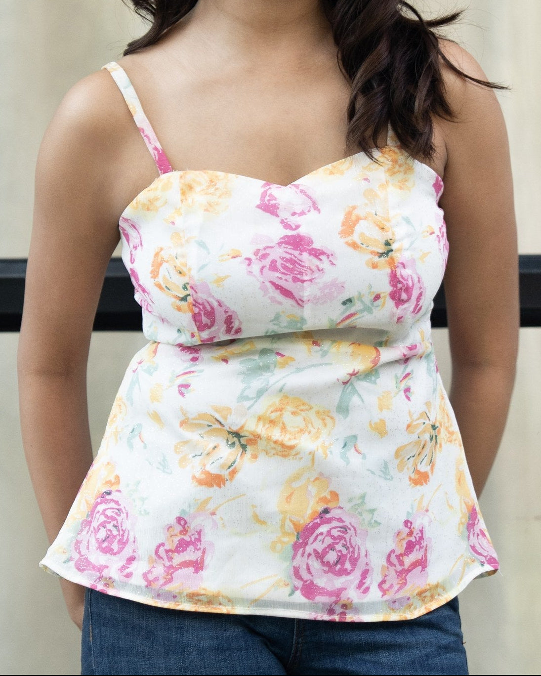 A woman smiling and wearing a white spaghetti-strap peplum top with a pink, yellow, and orange floral print, paired with blue jeans, set against a neutral beige background.